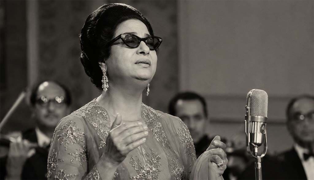 صورة واقعية بالأبيض والأسود للفنانة أم كلثوم أثناء الغناء على المسرح وسط فرقة موسيقية بطابع كلاسيكي قديم.